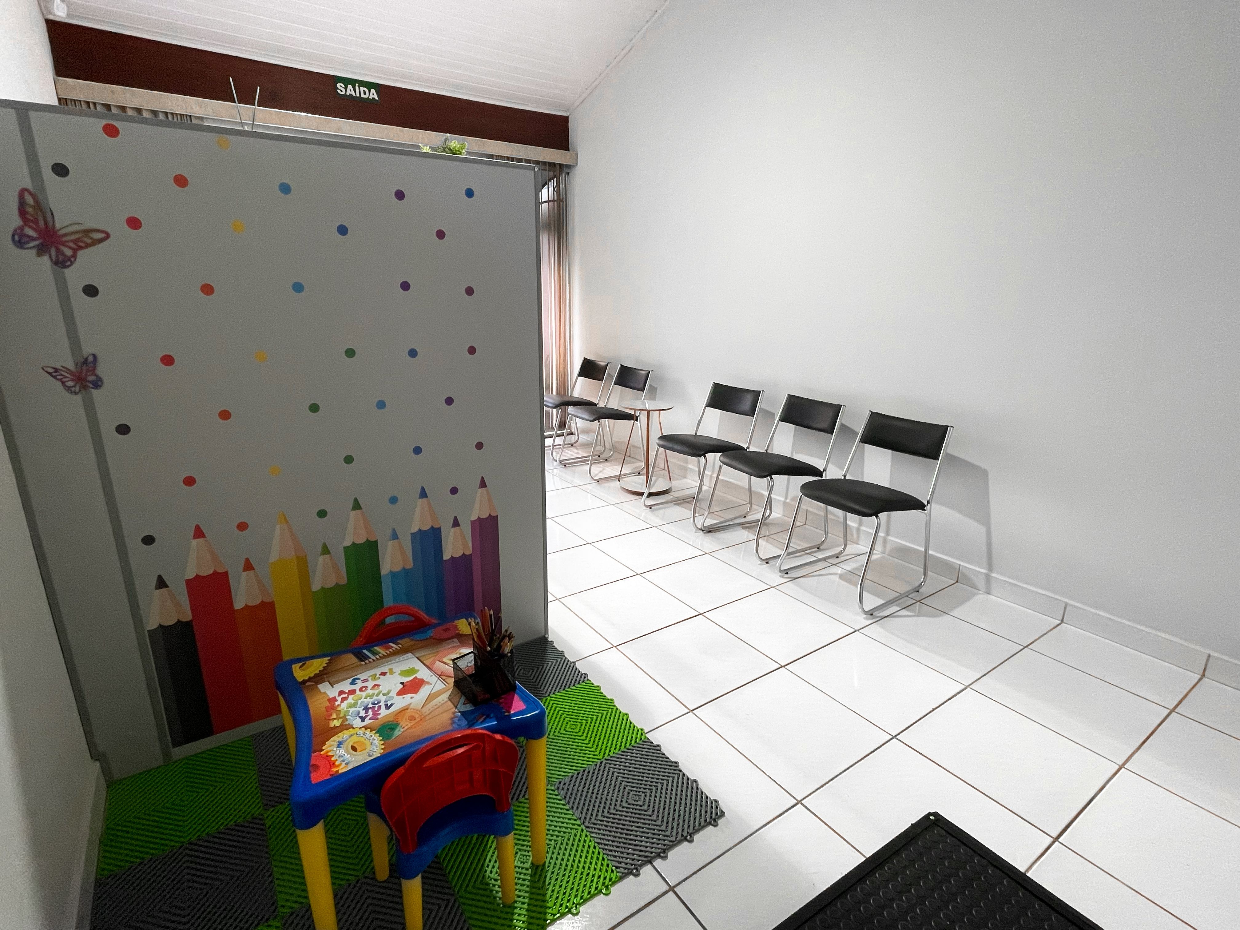 Espaço infantil com mesinha e cadeiras coloridas, ao lado das cadeiras da sala de espera.